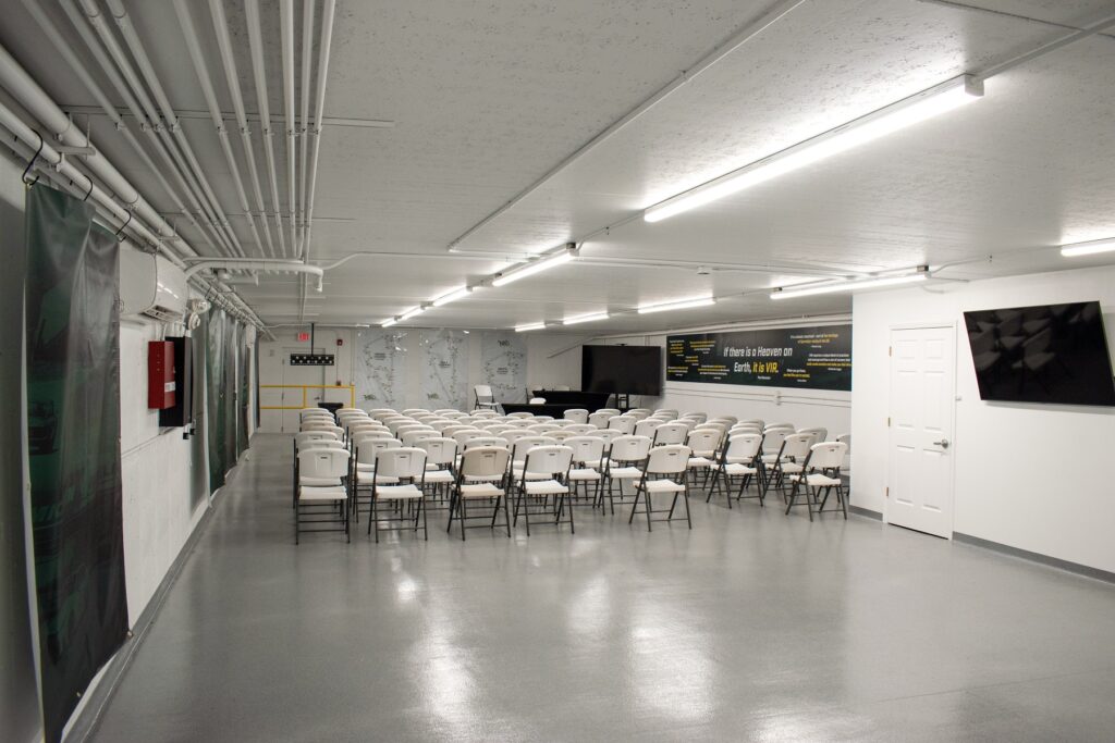 Drivers meeting room with white walls and white chairs with a green banner stating the quote "if there is a heaven on earth, it is VIR." in white and yellow writing among other smaller quotes. Large tv in the back of the photo with a table and track maps hanging on the wall