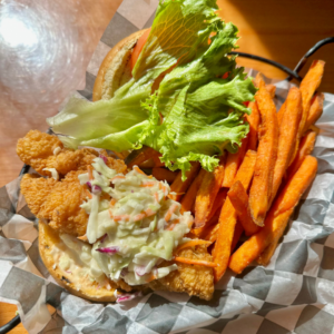 fried fish topped with cole slaw on a hamburger bun with lettuce and tomato with a side of sweet potato fries in a serving basket