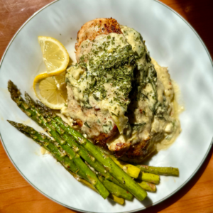 Baked chicken covered in a white sauce topped with herbs with a yellow lemon slice and green asparagus on the side on a white circle plate