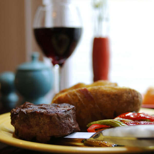 elegant photo of a steak, potatoes, and peppers on a dining plate with a silver knife and a glass of red wine in the background