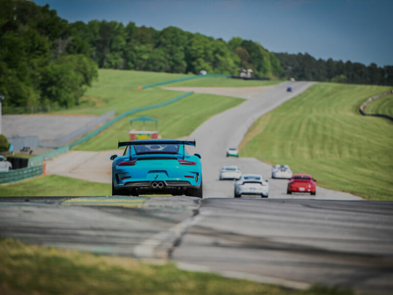 The back of a Miami blue Porsche GT3RS with black spoiler driving down the backstretch of VIR with other sports cars in the back ground of the image
