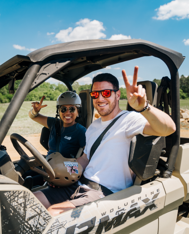 1 male and 1 female sitting in tan Yamaha Wolverine RMax ATV side by side holding up the peace sign