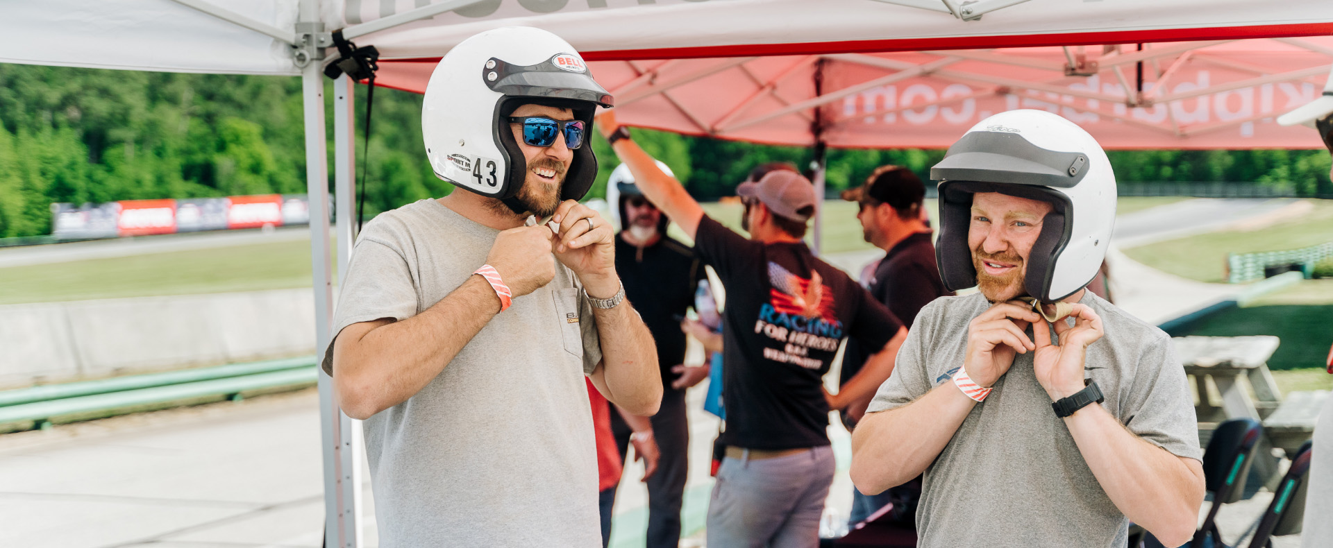 2 males buckling a white helmet chin straps waiting for a ride along on the race track